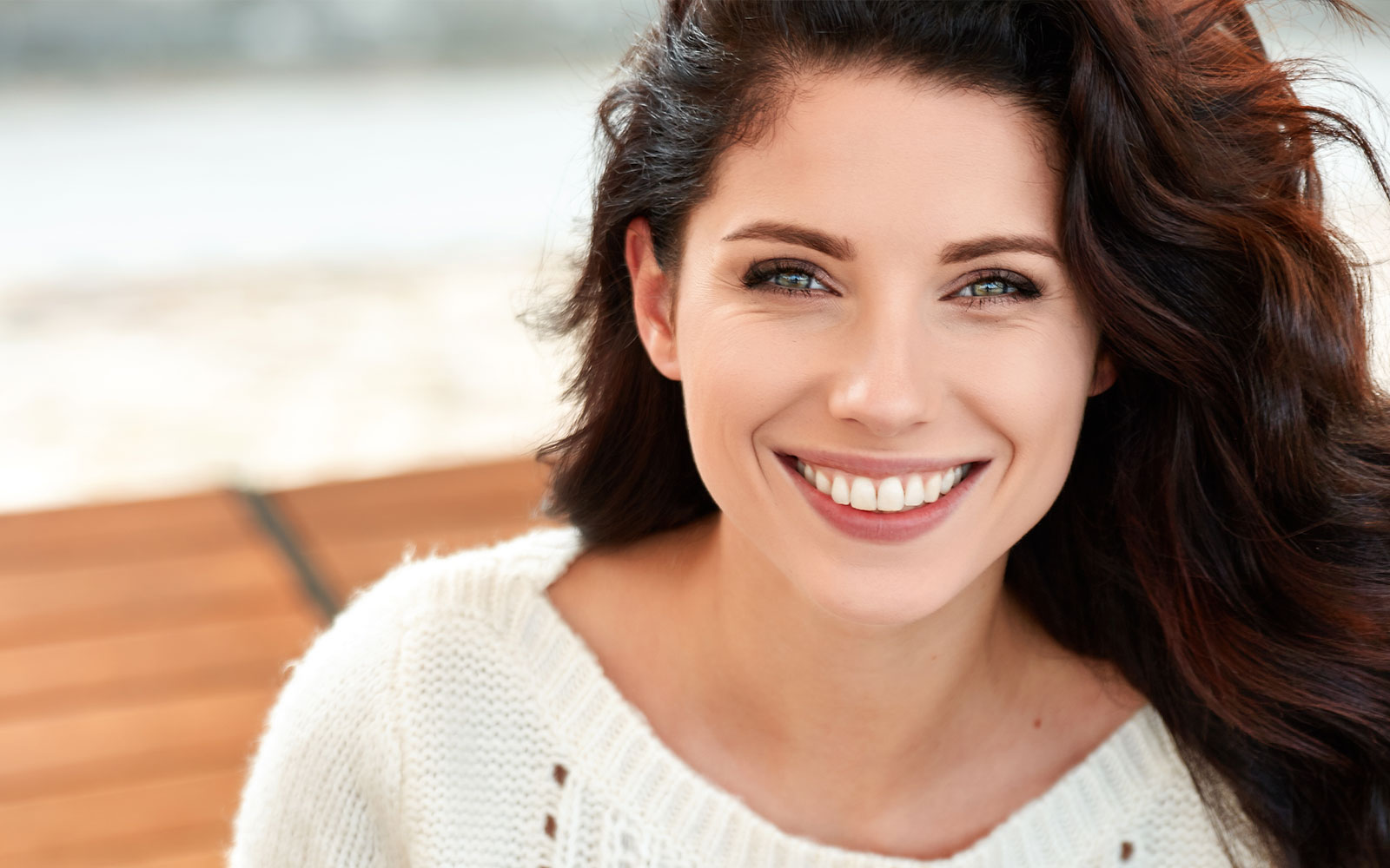 The image features a smiling woman with long hair, wearing a white top, set against a blurred background that appears to be an outdoor setting during daylight.