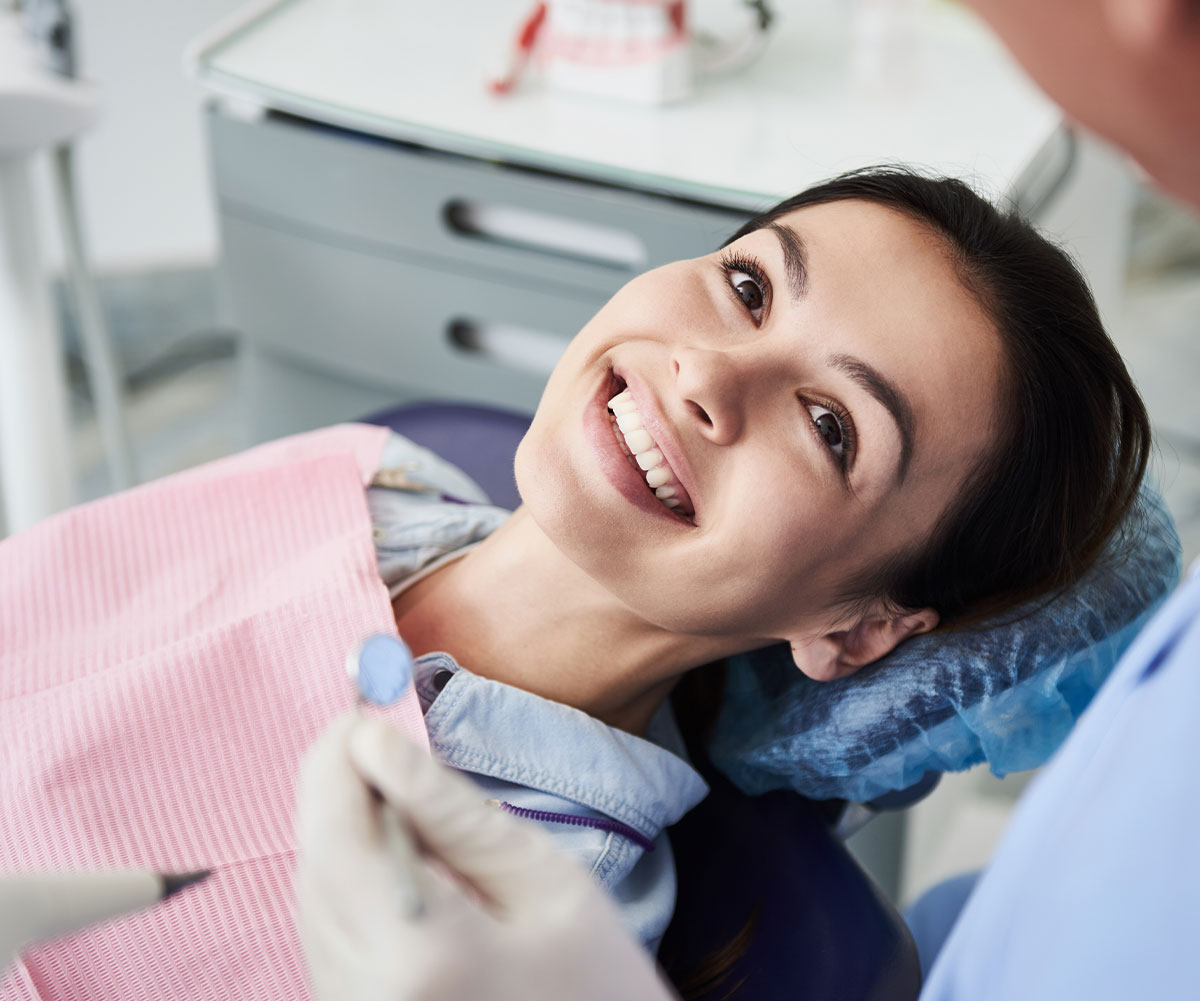 The image shows a woman sitting in a dental chair with a smile on her face, receiving dental care from a dentist who appears to be performing a procedure, with visible dental tools and equipment around them.