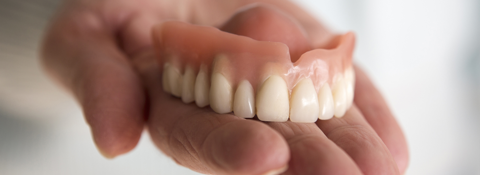 The image shows a person s hand holding a set of dentures with visible teeth in an indoor setting.