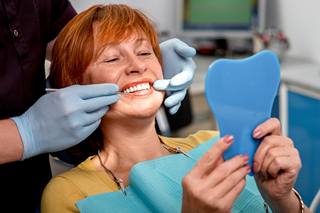 A woman wearing a surgical mask is holding up a blue dental model while seated in a dental chair, smiling as she looks at her reflection in a mirror.